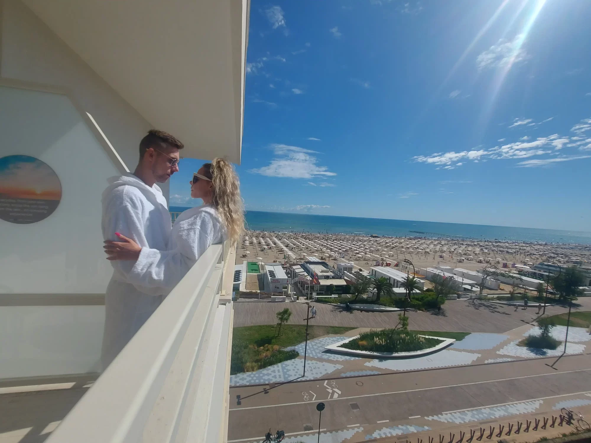 A couple in white robes stands on a balcony overlooking a beach under a sunny sky.