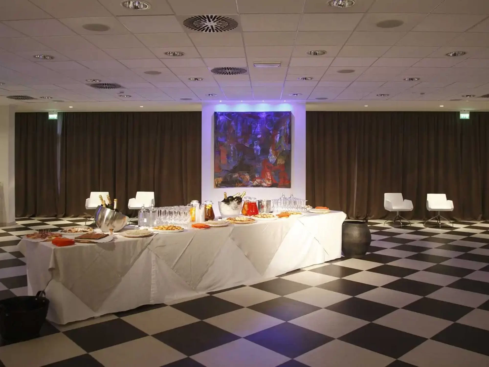 A banquet table with food in a checkered floor room with a colorful painting on the wall.